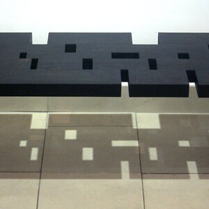 May include: A black wooden bench with a unique, geometric design. The bench has a series of cutouts in the top surface, creating a pattern of squares and rectangles.