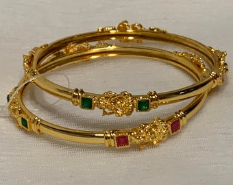 May include: Two gold bangles with intricate designs and green and red gemstones. The bangles are stacked on top of each other.