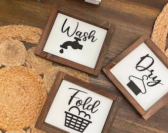 May include: Three wooden signs with black lettering and icons. The signs read "Wash" with a faucet icon, "Dry" with a hanger icon, and "Fold" with a basket icon.