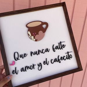 May include: A white wooden sign with a brown frame, featuring a pink heart and a brown coffee cup with pink and white floral designs. The sign reads "Que nunca falte el amor y el cafecito".
