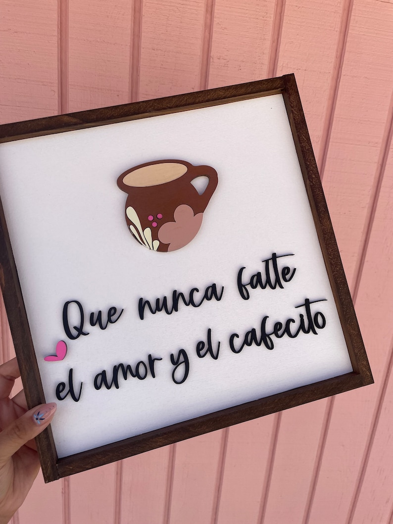 May include: A white wooden sign with a brown frame, featuring a pink heart and a brown coffee cup with pink and white floral designs. The sign reads "Que nunca falte el amor y el cafecito".