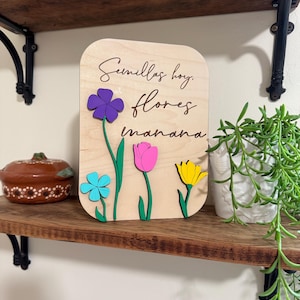 May include: Wooden sign with the text "Semillas hoy, flores manana" and colorful 3D flowers. The flowers include purple, blue, pink, and yellow. The sign is displayed on a wooden shelf with other decorative items.