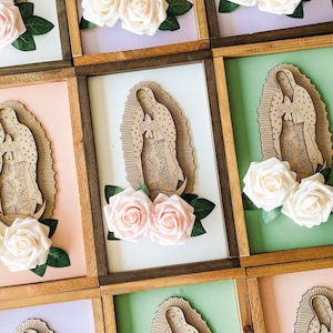 May include: A set of eight framed wood wall art pieces featuring a laser-cut image of the Virgin Mary with a floral design. Each frame has a different colored background, including pink, green, purple, and white. The frames are decorated with two white or pink artificial roses.