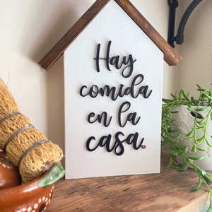 May include: White wooden house-shaped sign with black lettering that reads "Hay comida en la casa" with a small gold heart.