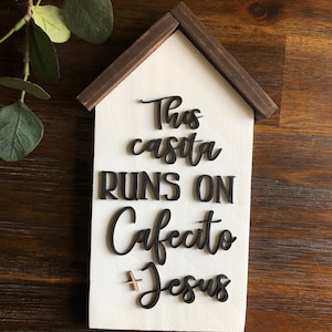 May include: A white wooden sign shaped like a house with a brown roof. The sign says "This casita runs on cafecito + Jesus".