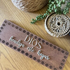 Puede incluir: Un letrero rectangular de madera con las palabras "DIOS bendiga este hogar" en escritura blanca. El letrero tiene un borde floral decorativo y est&aacute; colocado sobre una superficie de madera. Cestas tejidas y vegetaci&oacute;n en el fondo.