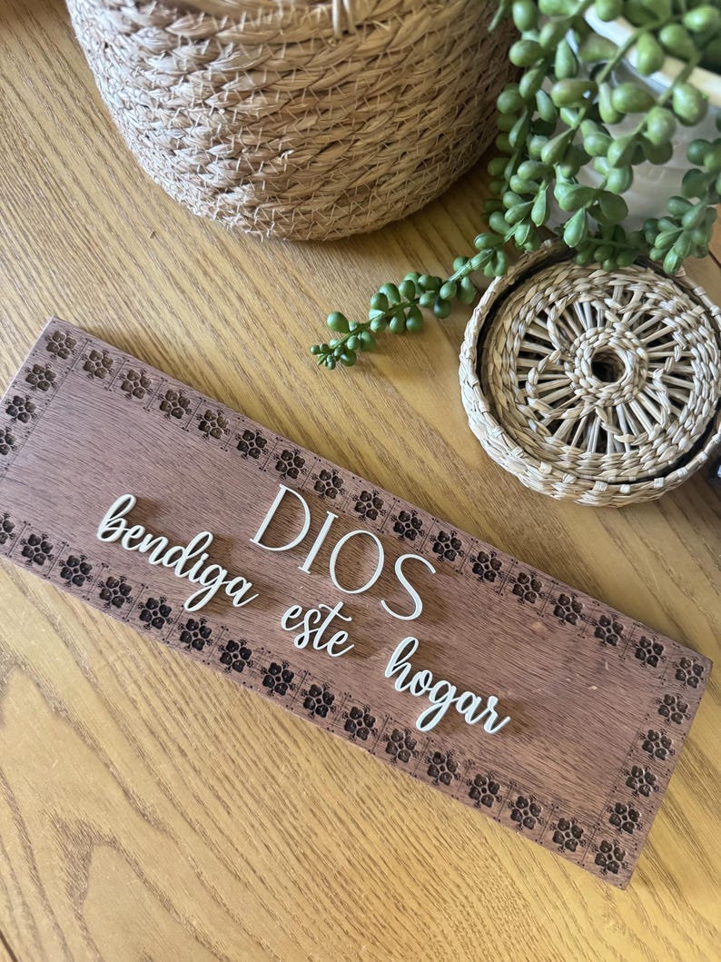 Puede incluir: Un letrero rectangular de madera con las palabras "DIOS bendiga este hogar" en escritura blanca. El letrero tiene un borde floral decorativo y est&aacute; colocado sobre una superficie de madera. Cestas tejidas y vegetaci&oacute;n en el fondo.