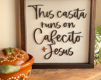 May include: A wooden sign with a white background and black lettering that reads "This casita runs on Cafecito + Jesus". The sign is framed in dark wood and is sitting on a wooden shelf.