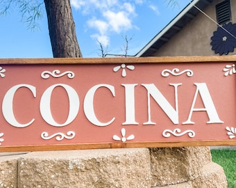 May include: A wooden sign with a brown frame and a pink background. The word "COCINA" is written in white letters on the sign. The sign has decorative white swirls and flower designs.