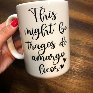 May include: White ceramic mug with black text that reads "This might be tragos de amargo licor" with two black hearts.