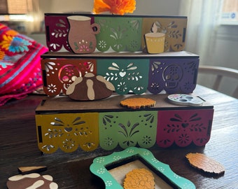 May include: Three-tiered wooden display with cut-out designs and colorful accents. The top tier features a marigold flower. Decorative cut-out shapes include a coffee mug, a volcano, and a skull. Additional wooden cut-outs are scattered around the display.