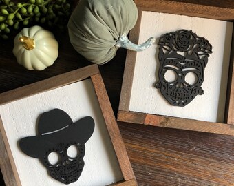 May include: Two framed wooden signs featuring black sugar skull designs. One skull is wearing a cowboy hat and the other is decorated with flowers.
