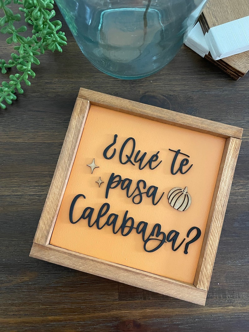 May include: A wooden sign with an orange background and black lettering that reads "&iquest;Qu&eacute; te pasa Calabaza?" with a small wooden pumpkin cutout.