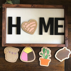 May include: Wooden sign with the word "HOME" in black letters. The letters are attached to a white background. Below the sign are five wooden cutouts: a brown pitcher with pink flowers, a pink, yellow, and brown candy, a green cactus in an orange pot, and a brown pig.