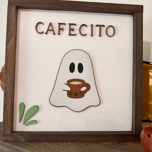 May include: A wooden framed sign with the word "Cafecito" in brown letters. A white ghost with black eyes holds a brown coffee mug with white polka dots.