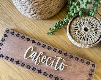 May include: A wooden sign with the word "Cafecito" in white script. The sign is bordered with a floral pattern and rests on a wooden surface. Woven baskets and a small plant are in the background.