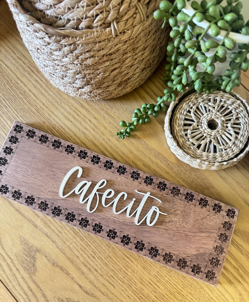Puede incluir: Un letrero de madera con la palabra "Cafecito" en escritura blanca. El letrero est&aacute; bordeado con un patr&oacute;n floral y descansa sobre una superficie de madera. Cestas tejidas y una peque&ntilde;a planta est&aacute;n en el fondo.