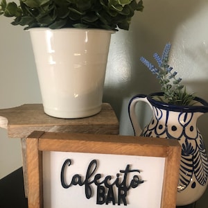 May include: A wooden sign with the words "Cafecito Bar" in black lettering. The sign is sitting on a wooden shelf with a white planter and a blue and white ceramic pitcher.