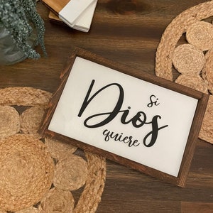 May include: A framed sign with the words "Dios si quiere" in black script. The sign has a white background and a dark brown wooden frame. Coasters and a decorative rug are also visible.