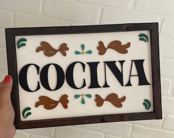 May include: A wooden sign with the word "COCINA" in black letters, surrounded by brown and teal decorative elements. The sign has a dark brown frame.