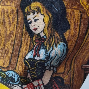 Puede incluir: Una ilustraci&oacute;n vintage de una mujer con un vestido azul y blanco con una cinta roja. Est&aacute; sosteniendo un plato con un dise&ntilde;o azul y blanco. El fondo es un panel de madera marr&oacute;n.