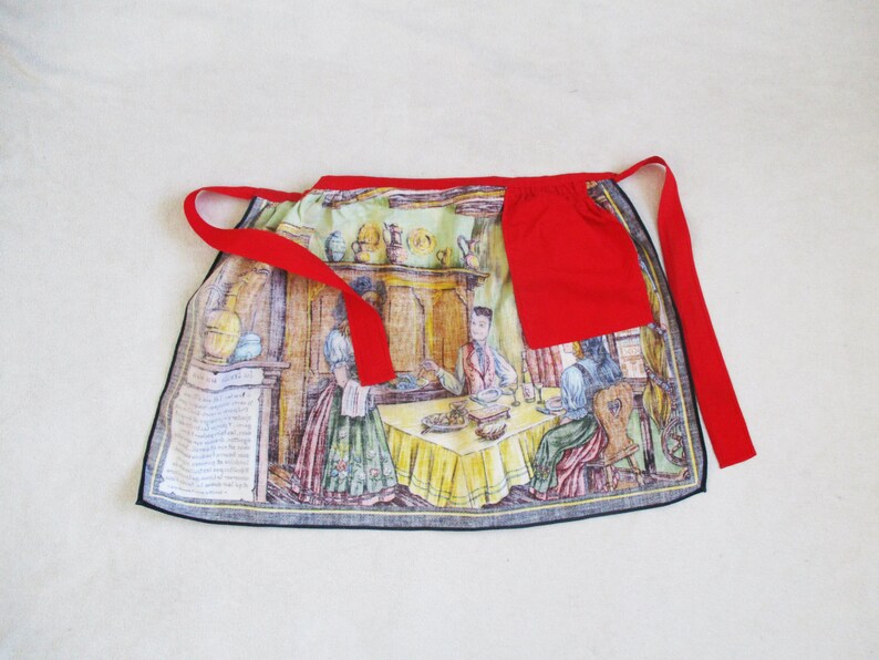 Puede incluir: Un delantal de cocina rojo y blanco con una ilustraci&oacute;n vintage de personas reunidas alrededor de una mesa. El delantal tiene un bolsillo rojo en la parte delantera y cintas rojas.