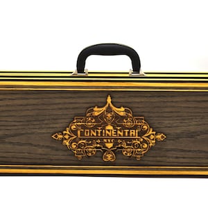May include: A rectangular wooden carrying case with a dark gray wood grain finish and yellow-gold trim. The center features an ornate laser-cut emblem with the text "CONTINENTAL NYC". It includes a black top handle and two black metal latches.