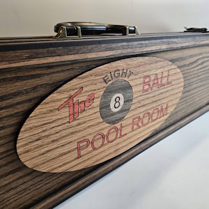 May include: A rectangular wooden case with a dark stained finish and a black handle. An oval wooden plaque is attached to the front, featuring the engraved text "The EIGHT BALL POOL ROOM" and a graphic of an eight ball in the center.