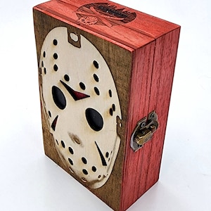 May include: A wooden box with a red exterior and a light-colored front featuring a cutout of a hockey mask. The box has a metal clasp and a dark interior. The top of the box has a carved design.