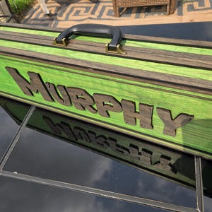 May include: A rectangular wooden carrying case with a bright green finish and dark brown wood grain accents. The name "MURPHY" is displayed in large, dark brown, raised wooden letters on the front. It features a black handle and metal latches.