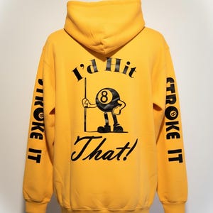 May include: Mustard yellow hoodie with black text and graphics. The back features a cartoon eight-ball character holding a pool cue, with the words "I'd Hit That!" The sleeves say "Stroke It".
