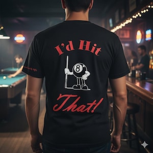 May include: Black t-shirt with the text "I'd Hit That!" in red, along with a cartoon eight-ball character holding a pool cue. The shirt is worn by a person in a dimly lit bar setting.