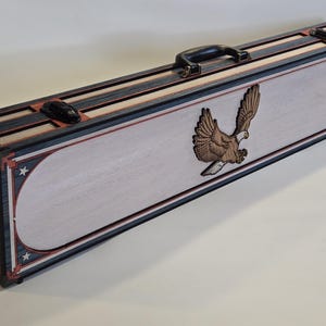 May include: A rectangular wooden case with a handle and a decorative eagle. The case features a light-colored central panel with a carved eagle, framed by red, white, and blue accents. The ends display a flag design.