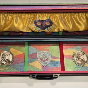 May include: Open wooden box with a colorful geometric design. The interior features a gold satin lining and a Majora's Mask emblem. Additional emblems include a shield and two portraits. The box is trimmed in red and black.