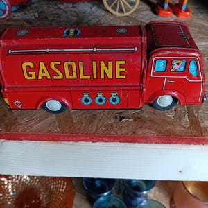 Könnte beinhalten: Ein roter Vintage-Spielzeug-Tanklaster aus Blech mit dem Wort GASOLINE in gelben Buchstaben an der Seite, blauen kreisförmigen Ventildetails und einem durch das Fenster sichtbaren Fahrer, mit schwarzen Rädern und einer abgenutzten Metalloberfläche.