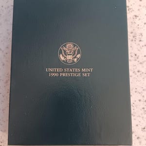 Könnte beinhalten: Eine dunkelgrüne rechteckige Präsentationsbox für ein United States Mint 1990 Prestige Set. Der Deckel zeigt ein goldenes kreisförmiges Siegel der United States Mint über dem Text UNITED STATES MINT 1990 PRESTIGE SET in goldener Schrift.