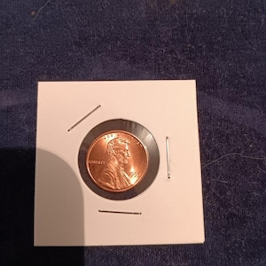 May include: A copper-coloured United States one-cent coin, known as a penny, encased in a square white cardboard holder. The coin features the profile of Abraham Lincoln, the word LIBERTY, the phrase IN GOD WE TRUST, and the year 1995 stamped on it.
