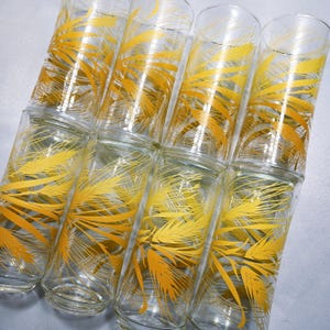 Könnte beinhalten: Ein Satz von acht transparenten Trinkgläsern mit einem gelben Weizenähren-Design. Die Gläser sind in zwei Reihen à vier Stück gestapelt und zeigen das sich wiederholende Muster. Das Design ist in einem warmen Gelbton gehalten.