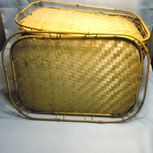 May include: Two rectangular bamboo serving trays. The trays have woven bamboo bases and curved bamboo frames. The trays are a light tan color. The trays are stacked on top of each other.