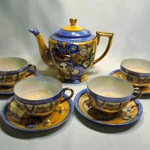 May include: A vintage tea set comprising a teapot and four teacups with saucers. The set is decorated in yellow, blue, and gold, with floral and abstract patterns. The teacups and saucers are arranged around the teapot.