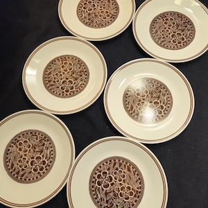 Könnte beinhalten: Sechs weiße Keramikplatten mit einem braunen Blumendesign in der Mitte. Jede Platte hat einen braunen Rand und ein detailliertes Blumenmuster. Die Teller sind auf einer dunklen Oberfläche angeordnet.