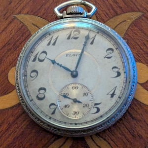 May include: A silver-toned Elgin pocket watch with a circular face, black Arabic numerals, and a sub-dial for seconds. The watch features a textured metal bezel, a winding crown at the top, and thin, dark-colored hands against a light dial.