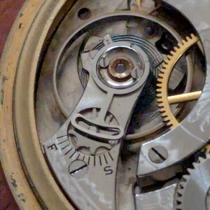 Puede incluir: Primer plano del mecanismo de un reloj antiguo, que muestra intrincados engranajes, tornillos y un volante. El mecanismo presenta un brazo regulador plateado con marcas 'F' y 'S', que indican ajuste r&aacute;pido y lento. La caja de lat&oacute;n del reloj es visible.