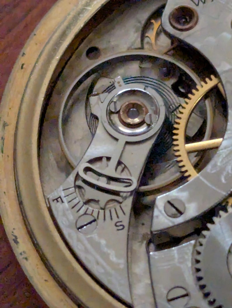 Puede incluir: Primer plano del mecanismo de un reloj antiguo, que muestra intrincados engranajes, tornillos y un volante. El mecanismo presenta un brazo regulador plateado con marcas 'F' y 'S', que indican ajuste r&aacute;pido y lento. La caja de lat&oacute;n del reloj es visible.
