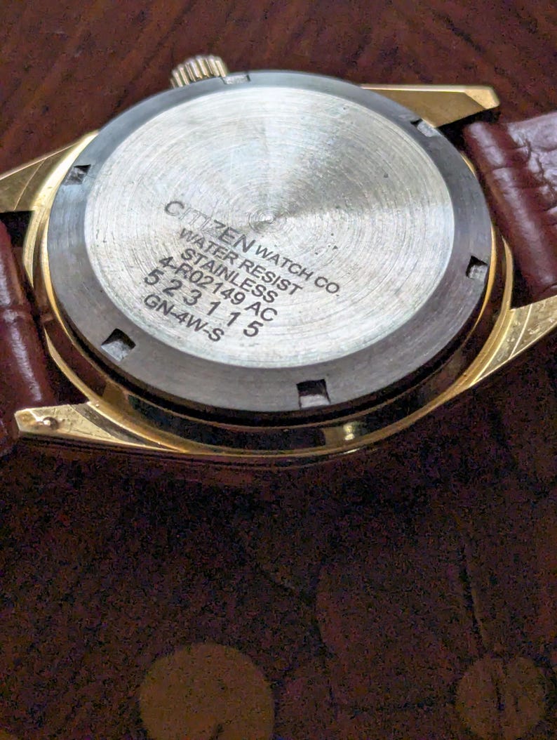 Pu&ograve; includere: Primo piano del retro di un orologio Citizen color oro con cinturino in pelle marrone. Il retro dell'orologio &egrave; in acciaio inossidabile e presenta il testo "CITIZEN WATCH CO. WATER RESIST STAINLESS 4-R02149 AC 523115 GN-4W-S".