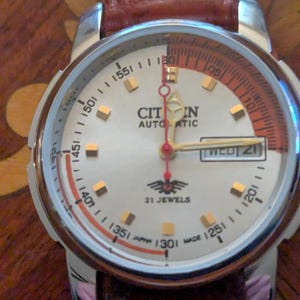 May include: A Citizen automatic wristwatch with a silver face, gold-toned markers, and a red and orange accent ring. The watch has a brown leather band and displays the day and date. The watch face reads "21 Jewels" and "Japan Made".