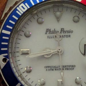 May include: A Philip Persio Illuminator wristwatch with a white dial, silver hands, and circular hour markers. The bezel features a blue and red color scheme with the number 20 visible. The dial text reads Philip Persio, Illuminator, Officially Certified, 3 ATM Water Proof, and Japan Movt.