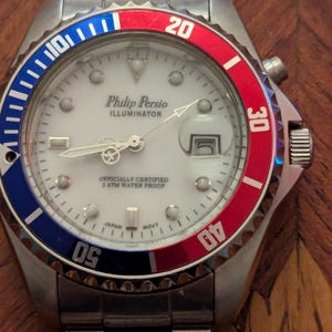 May include: A silver-tone analog wristwatch with a white dial, featuring the text Philip Persio ILLUMINATOR, OFFICIALLY CERTIFIED 3 ATM WATER PROOF, and JAPAN MOVT. The bezel is split into blue and red sections with white numerals.