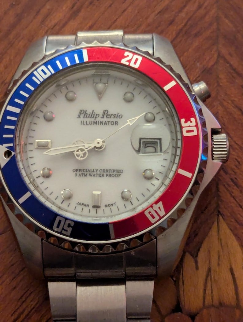 May include: A silver-tone analog wristwatch with a white dial, featuring the text Philip Persio ILLUMINATOR, OFFICIALLY CERTIFIED 3 ATM WATER PROOF, and JAPAN MOVT. The bezel is split into blue and red sections with white numerals.