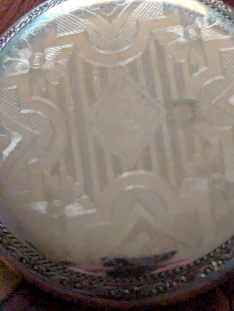 Puede incluir: Primer plano de la esfera de un reloj de bolsillo plateado con un intrincado dise&ntilde;o grabado. La esfera presenta una forma de diamante central rodeada de patrones florales y geom&eacute;tricos. El reloj tiene un borde detallado y texturizado.